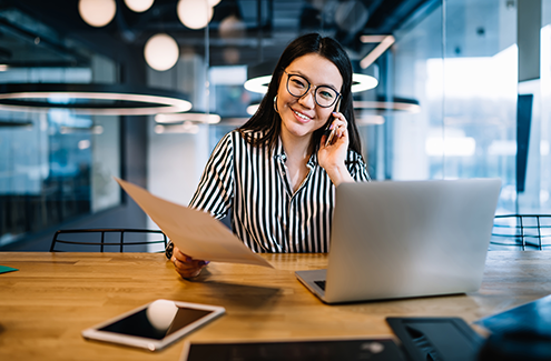 A female office worker sitting at a desk with a phone and computer, smiling.
